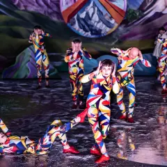 Artyści w kolorowych strojach tworzą syntezę sztuk w weselu, tańcząc na scenie z surrealistycznym tłem.