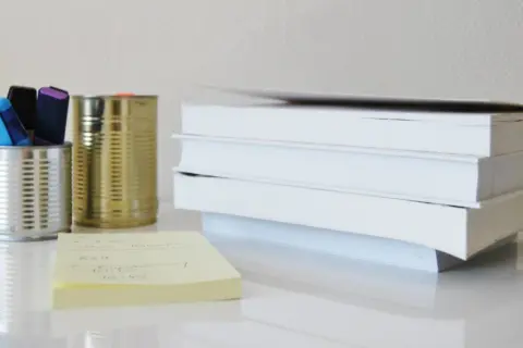 Na biurku stos białych książek, obok puszki z pisakami i żółta karteczka z notatkami, które mogą posłużyć jako akapity w rozprawce.