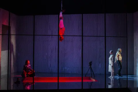 Scena z dramatu słowackiego: wisząca postać, siedząca kobieta w błyszczącym stroju, kamera i dwie stojące postacie.