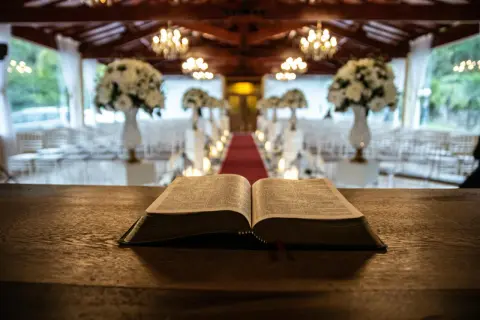 Otwarta Biblia na drewnianym stole, symbolizująca różne rodzaje ksiąg biblijnych. W tle udekorowana sala.