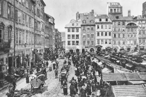 Tętniący życiem rynek, gdzie różne grupy społeczne w lalce handlują i spacerują wśród kamienic i straganów.