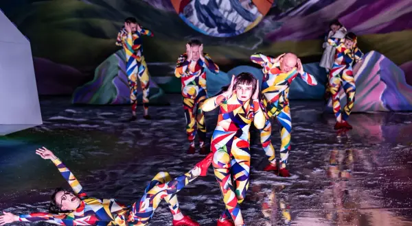 Artyści w kolorowych strojach tworzą syntezę sztuk w weselu, tańcząc na scenie z surrealistycznym tłem.